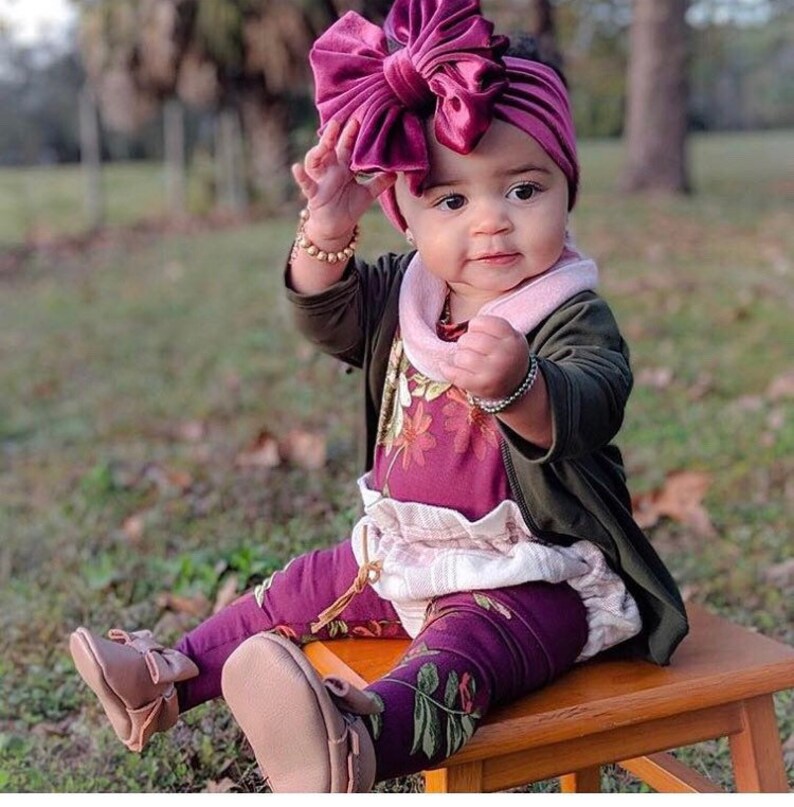 May include: A baby girl wearing a burgundy velvet headband with a large bow, a floral print top, a white ruffled skirt, and burgundy leggings with a floral print. She is wearing brown leather shoes and has a pink and white scarf around her neck.