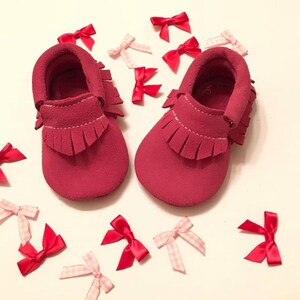 May include: A pair of red suede baby moccasins with fringe detailing. Scattered around the shoes are red and pink bows. The shoes are a vibrant shade of red, and the bows add a touch of whimsy. The moccasins are designed for infants.