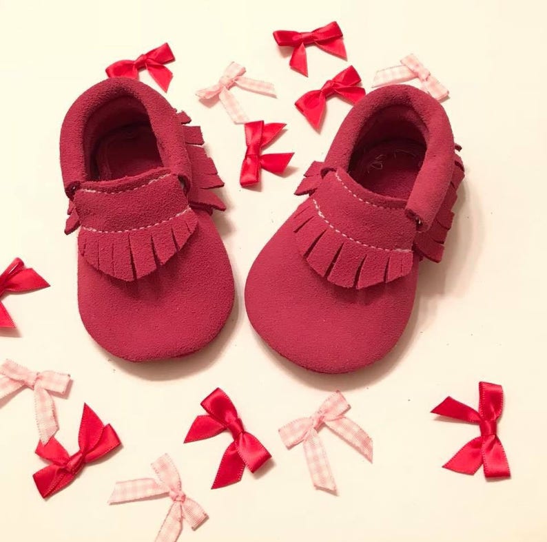May include: A pair of red suede baby moccasins with fringe detailing. Scattered around the shoes are red and pink bows. The shoes are a vibrant shade of red, and the bows add a touch of whimsy. The moccasins are designed for infants.