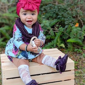 May include: A baby girl wearing a burgundy turban with a large bow, a floral print onesie, a brown scarf, white lace socks, and dark purple moccasins. She is holding a wooden teething ring with white beads.
