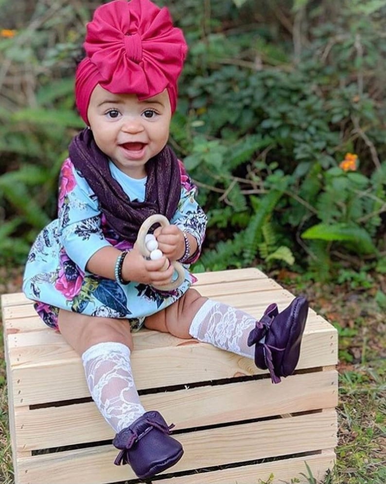 May include: A baby girl wearing a burgundy turban with a large bow, a floral print onesie, a brown scarf, white lace socks, and dark purple moccasins. She is holding a wooden teething ring with white beads.