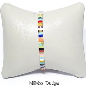 May include: A colorful bracelet made of small, square tiles. The tiles are arranged in a striped pattern with a variety of colors, including red, orange, yellow, green, blue, and purple. The bracelet is on a light gray pillow.