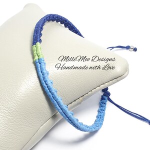 May include: A handmade macrame bracelet in shades of blue and green. The bracelet features a woven design with adjustable ties. The text "Milli Moo Designs Handmade with Love" is visible.