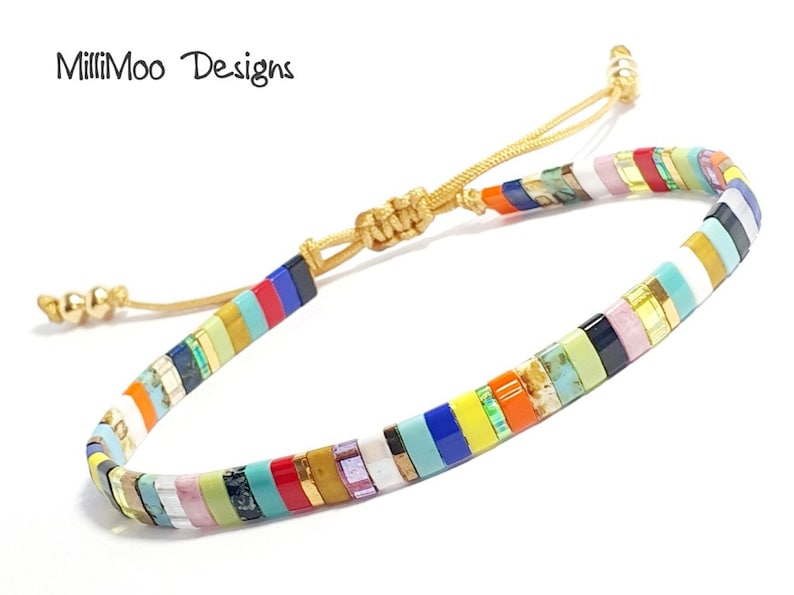 May include: Multicolored beaded bracelet with a gold cord closure. The beads are small and rectangular, and they are arranged in a striped pattern. The bracelet is adjustable.