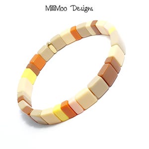 Peut inclure: Un bracelet coloré en carreaux carrés avec des carreaux beige, marron, orange et jaune. Le bracelet est en plastique et a un fermoir doré.