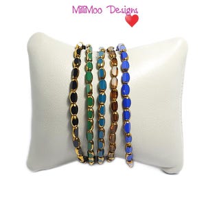 May include: Five bracelets made of gold-toned beads with different coloured glass inserts. The bracelets are arranged in a row on a white surface. The colours of the glass inserts are black, green, blue, brown, and blue.