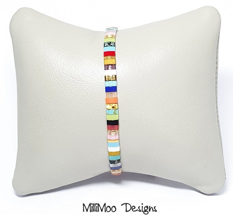 May include: A colorful bracelet made of small, square tiles. The tiles are arranged in a striped pattern with a variety of colors, including red, orange, yellow, green, blue, and purple. The bracelet is on a light gray pillow.