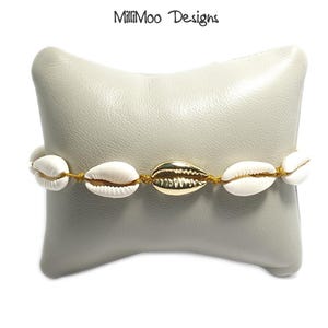 May include: A bracelet featuring white seashells and a gold-colored shell accent, strung on a gold-colored cord. The bracelet is displayed on a light gray pillow. The text "MilliMoo Designs" is in the upper right corner.