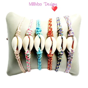 May include: Six handmade cowrie shell bracelets with colourful braided cords. The bracelets are displayed on a grey pillow. The cords are in various colours, including beige, pink, turquoise, red, purple, and a mix of colours. The brand name "MilliMoo Designs" is at the top.