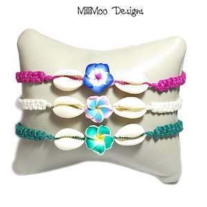 May include: Three handmade bracelets with white seashells and colourful flower accents. The bracelets have pink, white, and teal crochet bands. The top bracelet has a blue flower, the middle has a pink and blue flower, and the bottom has a teal flower.