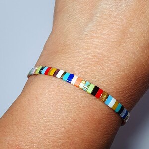 May include: A colorful beaded bracelet with a variety of colors, including red, orange, yellow, green, blue, and purple. The bracelet is made of small, square beads that are strung together on a thin, silver cord.