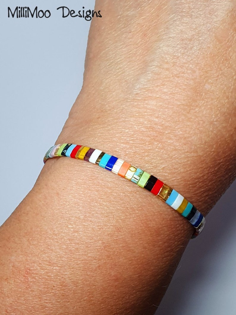 May include: A colorful beaded bracelet with a variety of colors, including red, orange, yellow, green, blue, and purple. The bracelet is made of small, square beads that are strung together on a thin, silver cord.