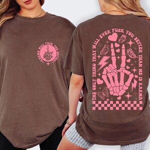 May include: Brown t-shirt with pink graphic designs. The front features a circular design with a hand giving the middle finger and the text "Karma will fuck you better". The back has a skeleton hand making a peace sign, butterflies, hearts, and the text "The only thing that will ever fuck you better than me is karma".