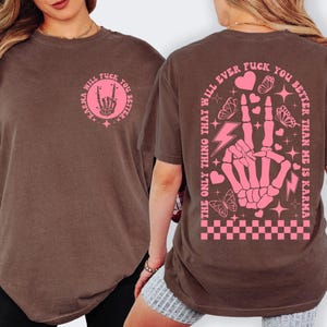 May include: Brown t-shirt with a pink graphic on the front and back. The front graphic says "Karma Will Fuck You Better" with a skeleton hand. The back graphic says "The Only Thing That Will Ever Fuck You Better Than Me Is Karma."