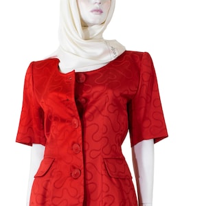 May include: A red skirt suit with a matching short-sleeved jacket. The jacket has a button-up front and two front pockets. The skirt is knee-length. The suit is displayed on a mannequin. The fabric has a repeating swirl pattern.