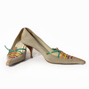 Peut inclure: Une paire d'escarpins en daim beige à bout pointu et talons aiguilles bas. L'avant de chaque chaussure présente un détail de laçage décoratif avec de fines brides brillantes vertes, orange, jaunes et violettes partant des côtés vers le centre.