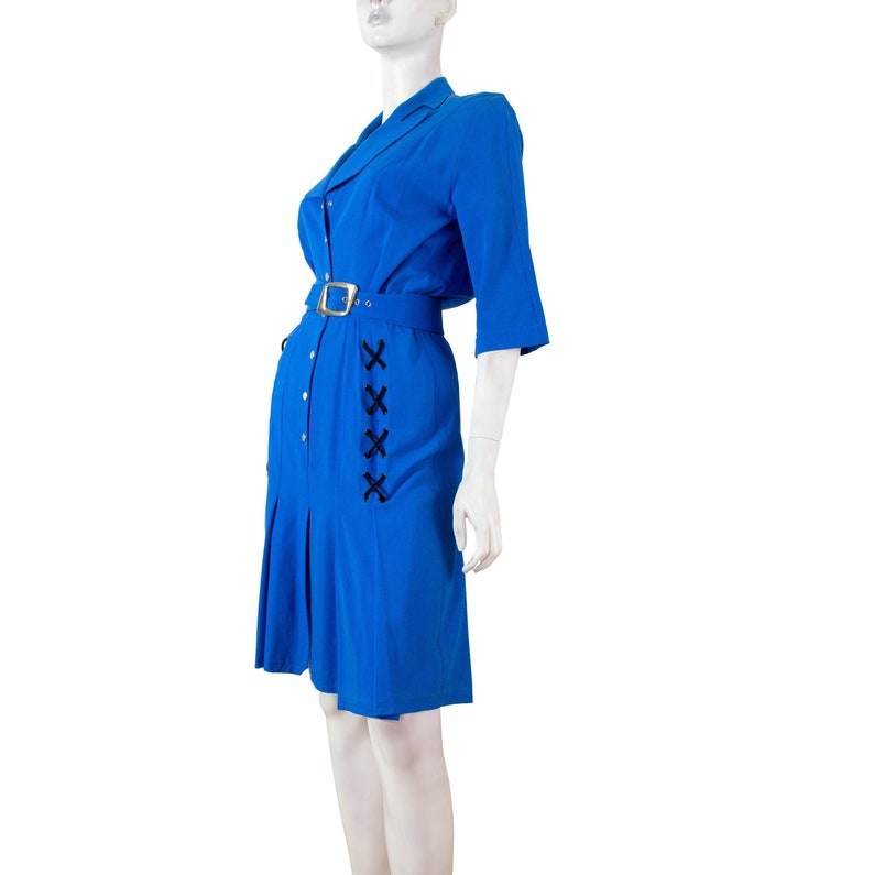 May include: A blue dress with a white collar and a black belt. The dress has a button-up front and a pleated skirt. There is a black lace-up detail on the side of the dress.