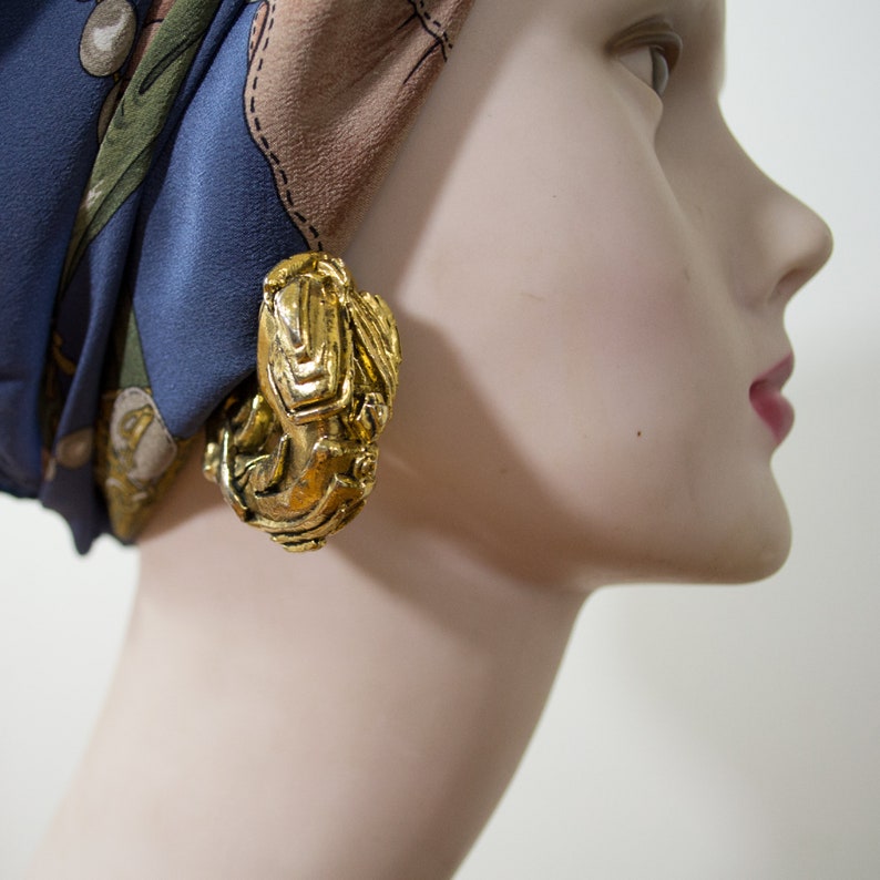 Op de afbeelding: Goudkleurige, getextureerde oorringen. De oorbellen hebben een sierlijk, gebeeldhouwd ontwerp. De oorbellen worden getoond op een mannequin met een blauw en bruin patroon sjaal. Een opvallend sieraad.
