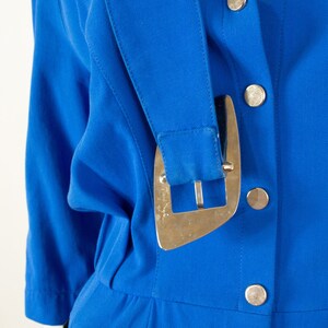 May include: A close-up of a blue jumpsuit with a gold buckle and silver buttons. The jumpsuit has a collar and long sleeves.