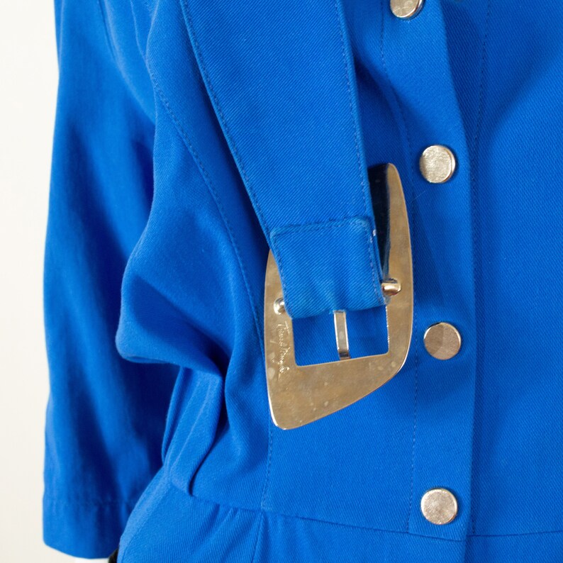 May include: A close-up of a blue jumpsuit with a gold buckle and silver buttons. The jumpsuit has a collar and long sleeves.
