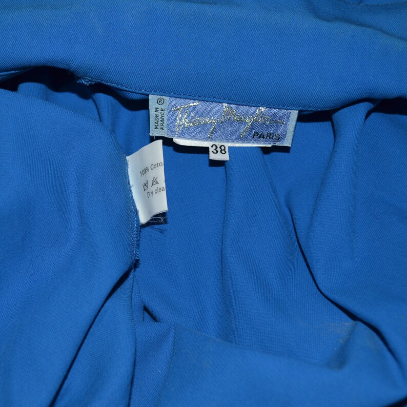 May include: A blue cotton shirt with a white tag that says "Made in France" and "100% Cotton" with a dry clean symbol. The tag also has the brand name "Thierry Mugler" and "Paris" printed on it. The shirt has a size tag that says "38".