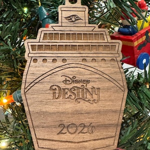 May include: Wooden Christmas ornament shaped like a cruise ship, with the words "Disney Destiny" and the year "2020" engraved on it. The ornament is hanging on a Christmas tree.