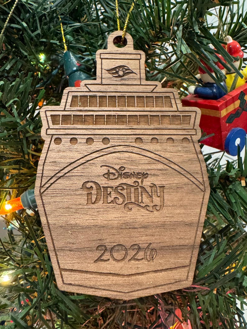 May include: Wooden Christmas ornament shaped like a cruise ship, with the words "Disney Destiny" and the year "2020" engraved on it. The ornament is hanging on a Christmas tree.