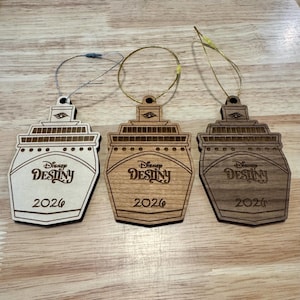 May include: Three wooden cruise ship ornaments with the text "Disney Destiny 2026". The ornaments are in shades of light, medium, and dark brown, each with a different hanging string.