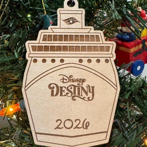 May include: Wooden Christmas ornament shaped like a cruise ship, featuring the words "Disney Destiny" and the year "2026". The ornament is hanging on a Christmas tree with green needles and colorful lights.