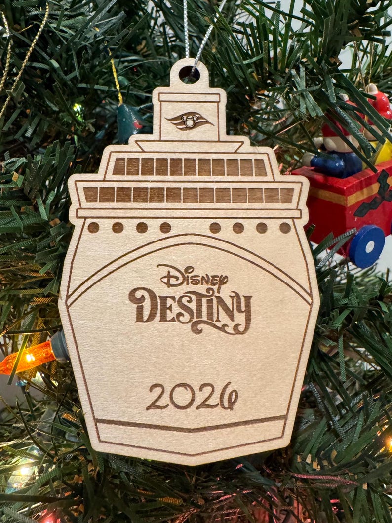 May include: Wooden Christmas ornament shaped like a cruise ship, featuring the words "Disney Destiny" and the year "2026". The ornament is hanging on a Christmas tree with green needles and colorful lights.