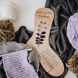 May include: A wooden sock knitting loom with a leaf design and the text "Knitted with love by Corrianne". There are balls of yarn in various colours, including white, purple, and brown.