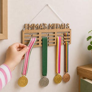 Pu&ograve; includere: Espositore di medaglie in legno con la scritta "EMMA'S MEDALS". L'espositore contiene diverse medaglie con nastri colorati. Una mano sta posizionando una medaglia con un nastro a righe rosa e bianco. Un'immagine incorniciata con un cuore rosa &egrave; sullo sfondo.