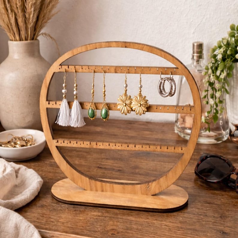 May include: A wooden earring holder with a circular design. The holder displays various earrings, including white tassel earrings, gold drop earrings with green stones, and gold flower earrings. The holder is made of light-colored wood and sits on a wooden surface.
