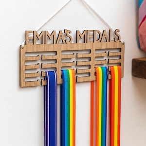 Pu&ograve; includere: Espositore di medaglie in legno con il testo "EMMA'S MEDALS" intagliato. Presenta fessure per nastri, con nastri blu, arcobaleno e arancioni appesi. Un nastro bianco viene utilizzato per appendere.