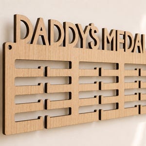 Pu&ograve; includere: Espositore di medaglie in legno con le parole "DADDY'S MEDALS" intagliate nella parte superiore. L'espositore ha pi&ugrave; fessure orizzontali per appendere le medaglie. Il legno &egrave; di colore marrone chiaro.