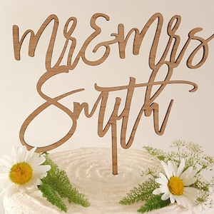 May include: Wooden cake topper with the words "Mr & Mrs Smith" in a cursive font. The topper is on a white cake with green leaves and white daisies.