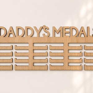 Pu&ograve; includere: Espositore di medaglie in legno chiaro con la scritta "DADDY'S MEDALS" intagliata nella parte superiore. L'espositore ha tre file di supporti per medaglie. Il legno &egrave; marrone chiaro e presenta venature naturali.