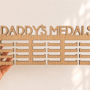 Pu&ograve; includere: Un espositore di medaglie in legno con le parole "DADDY'S MEDALS" ritagliate nella parte superiore. Il display ha tre file di fessure per appendere le medaglie. Il display &egrave; tenuto da una mano.