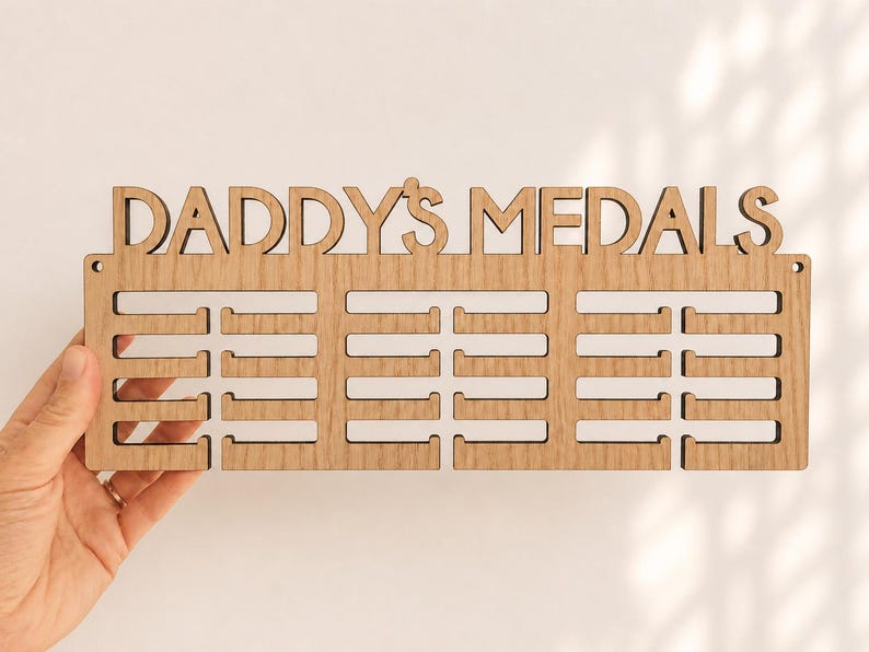 Pu&ograve; includere: Un espositore di medaglie in legno con le parole "DADDY'S MEDALS" ritagliate nella parte superiore. Il display ha tre file di fessure per appendere le medaglie. Il display &egrave; tenuto da una mano.