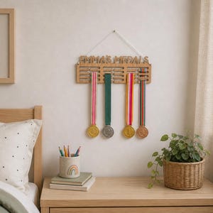 Pu&ograve; includere: Un espositore di medaglie in legno con il nome "EMMA'S MEDALS" inciso. Espone quattro medaglie con nastri colorati. Una stampa incorniciata con la scritta "be your best self" &egrave; appesa al muro. Una piccola pianta in vaso &egrave; su un comodino di legno.