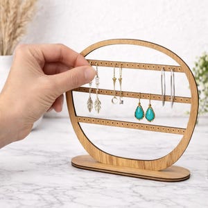 May include: A wooden earring display stand with a circular design. The stand holds various earrings, including silver hoops, gold drop earrings, and turquoise teardrop earrings. The stand is made of light-colored wood and has a base for stability.