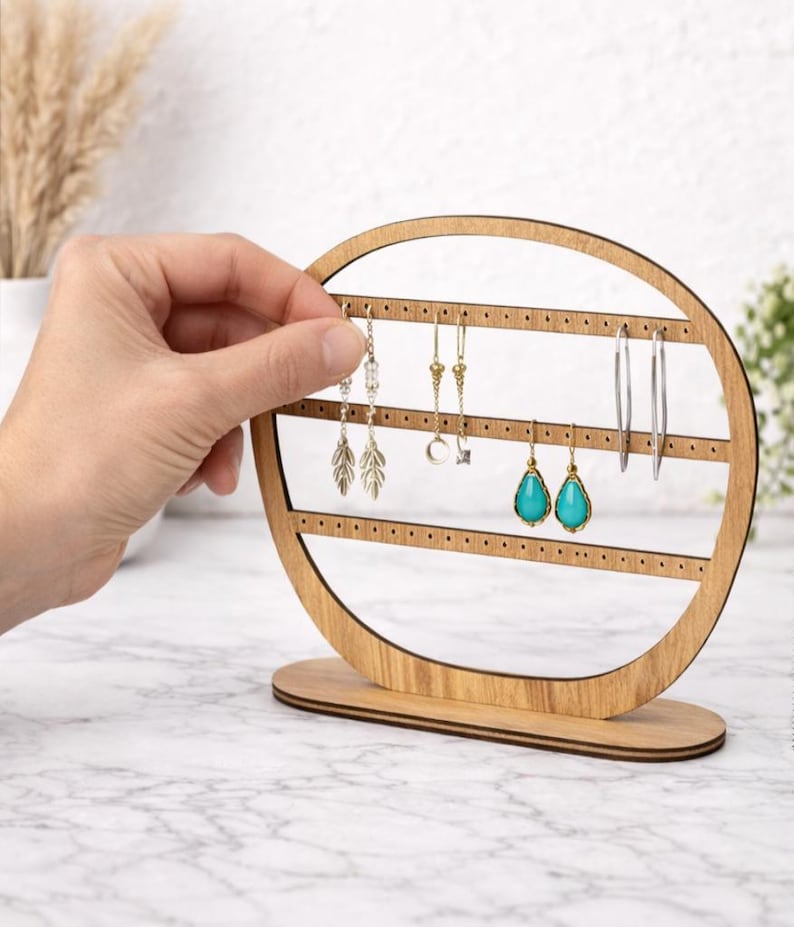 May include: A wooden earring display stand with a circular design. The stand holds various earrings, including silver hoops, gold drop earrings, and turquoise teardrop earrings. The stand is made of light-colored wood and has a base for stability.