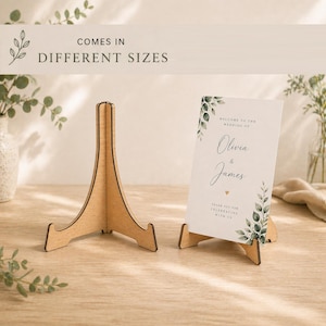 May include: A wooden easel stand next to a wedding sign. The sign says "Welcome to the Wedding of Olivia & James" and is decorated with green leaves. The text "Comes in Different Sizes" is at the top.