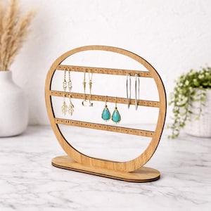 May include: A wooden earring display stand with a light brown finish. The stand has an oval shape with multiple horizontal rows of small holes to hold earrings. Several pairs of earrings, including silver hoops and turquoise teardrop earrings, are displayed.