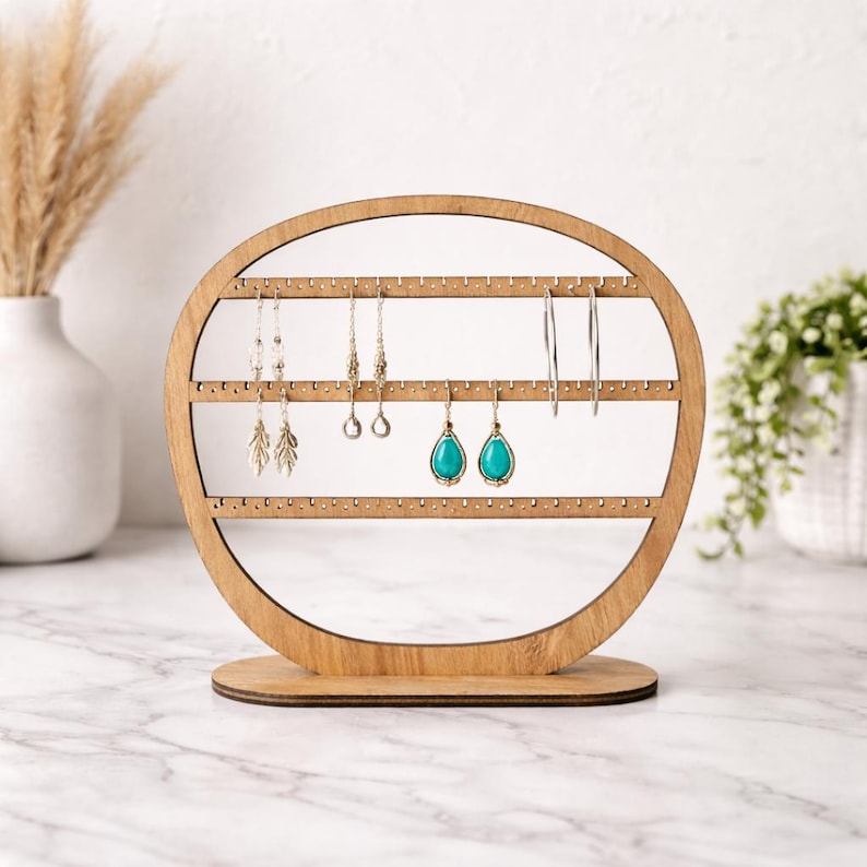 May include: A wooden earring holder with an oval frame and three horizontal rows of small holes for displaying earrings. Several pairs of earrings, including silver hoops and turquoise teardrop earrings, are displayed on the holder.