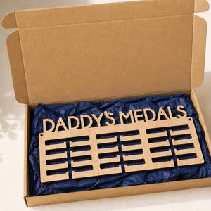 Pu&ograve; includere: Un espositore di medaglie in legno con la scritta "DADDY'S MEDALS". Presenta tre file di fessure per medaglie ed &egrave; presentato in una scatola di cartone aperta con carta velina blu.