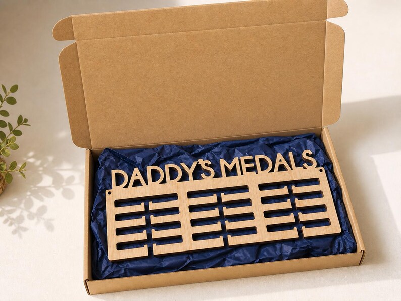 Pu&ograve; includere: Un espositore di medaglie in legno con la scritta "DADDY'S MEDALS". Presenta tre file di fessure per medaglie ed &egrave; presentato in una scatola di cartone aperta con carta velina blu.