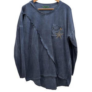 May include: A long-sleeved, oversized, blue-grey top with a unique asymmetrical design. Features a small pocket with a star embellishment. The top is hanging on a wooden hanger.