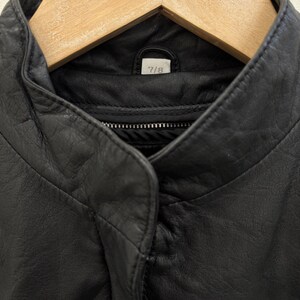 以下が含まれることがあります： 黒い革のジャケットの襟のクローズアップ。 ジャケットはスタンドカラーとジッパー開閉です。 タグには「7/8」と書かれています。 ジャケットは木製のハンガーに掛かっています。