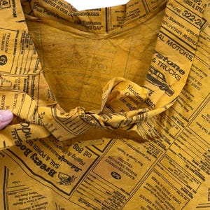 Może przedstawiać: Pognieciona ż&oacute;łta papierowa torba z nadrukiem gazety. Torba jest otwarta, odsłaniając wnętrze. Nadruk gazety zawiera r&oacute;żne reklamy i tekst. Torba jest prawdopodobnie używana do pakowania lub przenoszenia przedmiot&oacute;w.